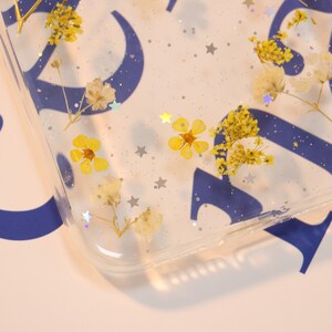 May include: A clear phone case with dried flowers and silver glitter embedded in the resin. The flowers are yellow and white.