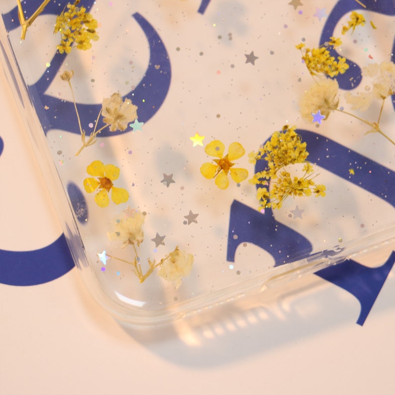 May include: A clear phone case with dried flowers and silver glitter embedded in the resin. The flowers are yellow and white.