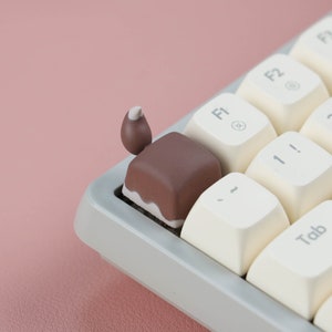 May include: A white and brown keyboard keycap shaped like a chocolate bar with a white swirl on top. The keycap is on a white keyboard with black keys.