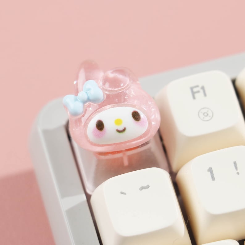 Puede incluir: Un tap&oacute;n de tecla rosa y blanco con un lazo azul. El personaje tiene una cara rosa con ojos marrones, una nariz amarilla y una boca rosa. El tap&oacute;n de tecla est&aacute; dise&ntilde;ado para encajar en un teclado mec&aacute;nico.