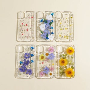 Könnte beinhalten: Sechs transparente Handyhüllen mit gepressten Blumen und Glitzer im Inneren. Die Hüllen zeigen verschiedene Blumenarrangements, darunter blaue, lila, gelbe und weiße Blumen.