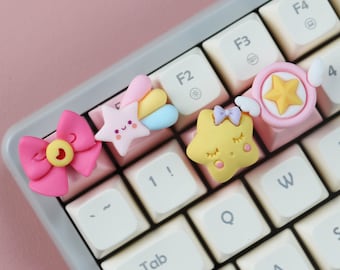 Könnte beinhalten: Vier bunte Tastenkappen mit niedlichen Comic-Designs: eine rosa Schleife mit einem Stern, ein gelber Stern mit geschlossenen Augen, ein Regenbogen-Sternschnuppen und ein rosa Zauberstab mit einem Stern. Die Tastenkappen befinden sich auf einer weißen Tastatur mit rosa Tasten.