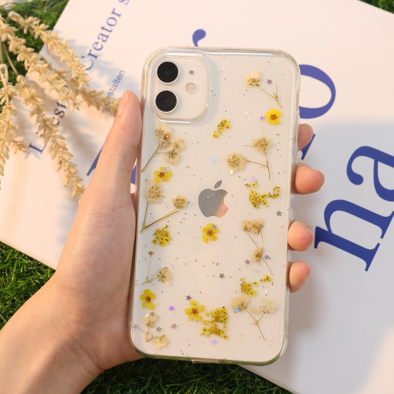 May include: A clear phone case with a white background and a design of pressed dried flowers and glitter. The flowers are yellow and white, and the glitter is silver.