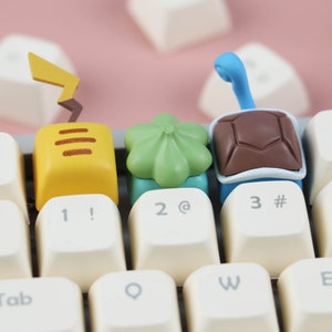 May include: Three colorful keycaps featuring a yellow Pikachu, a green Bulbasaur, and a blue Squirtle. The keycaps are on a white keyboard with the numbers 1, 2, and 3.