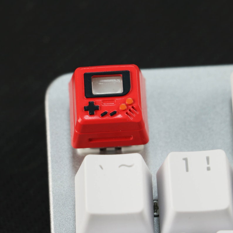 May include: A red keycap shaped like a retro video game console with a black screen, a black plus sign, and orange buttons. The keycap is on a white keyboard.