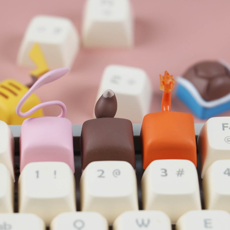May include: Three keycaps with different designs: a pink keycap with a curled tail, a brown keycap with a small brown bump, and an orange keycap with a flame-shaped top. The keycaps are on a white keyboard with black keys.