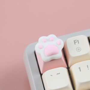 May include: A white keyboard keycap with a pink paw print design. The keycap is on a pink keyboard.