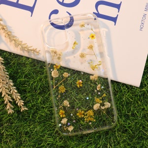May include: Clear phone case with dried flowers and glitter embedded inside. The flowers are white and yellow.