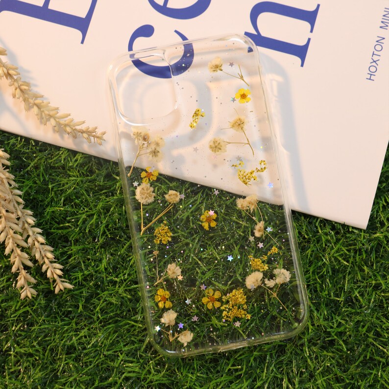 May include: Clear phone case with dried flowers and glitter embedded inside. The flowers are white and yellow.