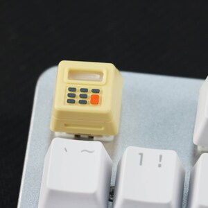 May include: A yellow plastic keycap with a calculator design. The keycap has a small screen, four buttons, and a red button. The keycap is on a white keyboard.