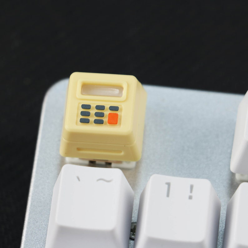 May include: A yellow plastic keycap with a calculator design. The keycap has a small screen, four buttons, and a red button. The keycap is on a white keyboard.