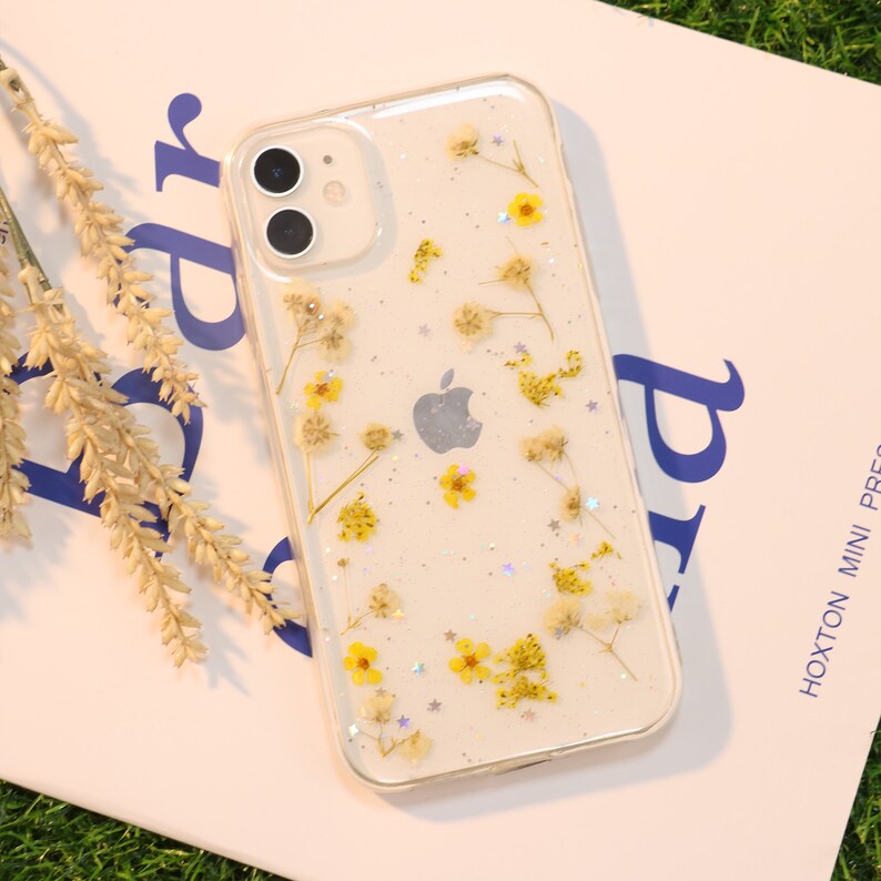 May include: A clear phone case with a white background and a design of pressed dried flowers and glitter. The phone case is for an iPhone.