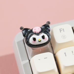 Puede incluir: Un tap&oacute;n de tecla negro y rosa Hello Kitty con una calavera en la parte superior. El tap&oacute;n de tecla est&aacute; en un teclado blanco.