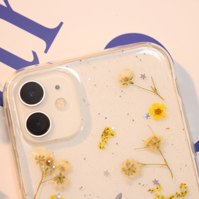 May include: A clear phone case with a white background and glitter.  The case has dried flowers and silver stars embedded in the plastic.