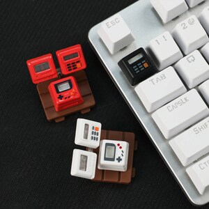 May include: A set of miniature keyboard keycaps featuring a calculator, a video game console, and a computer. The keycaps are arranged on a brown base and are displayed on a black surface.
