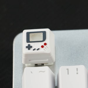 May include: White keyboard keycap with a black and gray Game Boy design. The keycap has a red and gray button design and a gray screen.