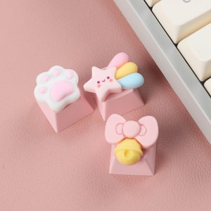 May include: Three pink keyboard keycaps with cute cartoon designs. One keycap features a white paw print, another has a pink star with a rainbow and a smiling face, and the last one has a pink bow with a yellow bell.