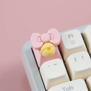 May include: A pink keyboard keycap with a bow and a yellow bell charm. The keycap is on a white keyboard.