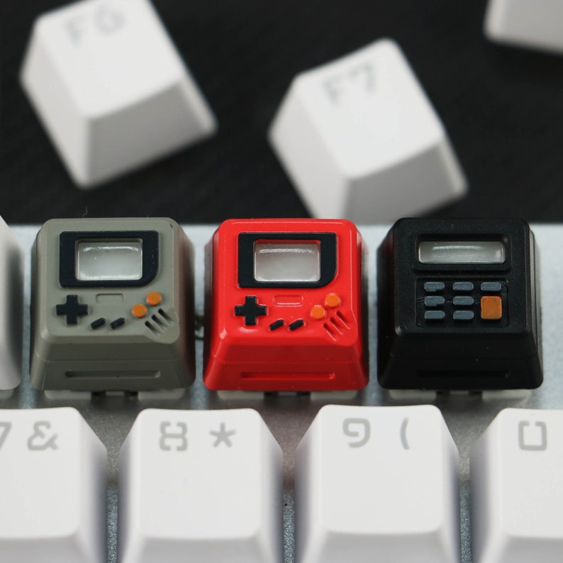 May include: Three miniature keyboard keycaps with a retro video game design. The keycaps are gray, red, and black with a pixelated screen, buttons, and joystick. The keycaps are on a white keyboard.