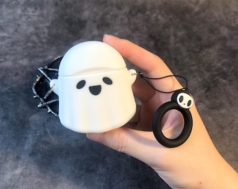 Puede incluir: Una funda blanca con forma de fantasma para auriculares inalámbricos con una cara sonriente negra. La funda tiene un cordón negro a juego y un anillo negro con un diseño de calavera. La funda se sostiene en una mano.