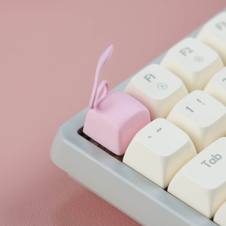 May include: A pink keyboard keycap with a unique design resembling a rabbit ear. The keycap is placed on a white keyboard with other keys visible, including "F1", "F2", "Tab", and "1!".