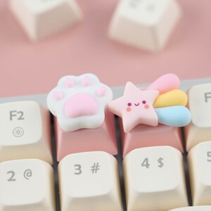 May include: Two pink and white keycaps with cute designs. One keycap features a paw print and the other a star with a rainbow tail. The keycaps are on a pink and white keyboard.