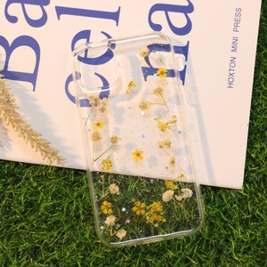 May include: A clear phone case with dried flowers and glitter embedded inside. The flowers are white and yellow, and the glitter is silver.
