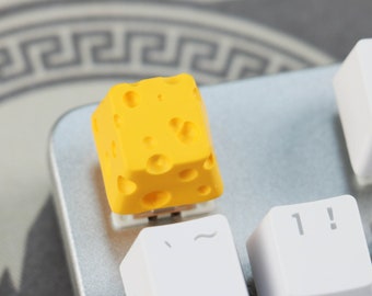 May include: A yellow cheese-shaped keycap for a mechanical keyboard. The keycap is textured to look like a block of Swiss cheese.