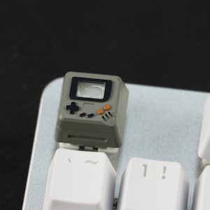 May include: A gray and black keycap with a retro video game console design. The keycap has a black screen, orange buttons, and a black directional pad.