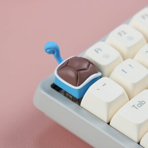 May include: A small, brown turtle-shaped keycap with a blue shell and a blue tail. The keycap is on a white keyboard with a white key labeled "Tab".