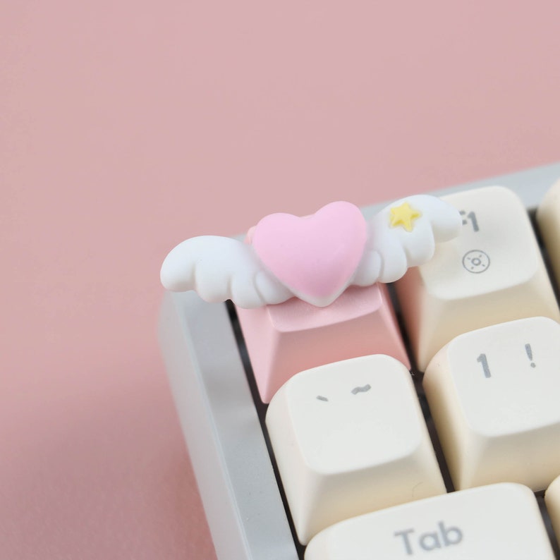 May include: A pink heart with white wings and a yellow star sits on top of a pink keyboard key. The key is labeled with a tilde symbol. The keyboard is white with black keys.