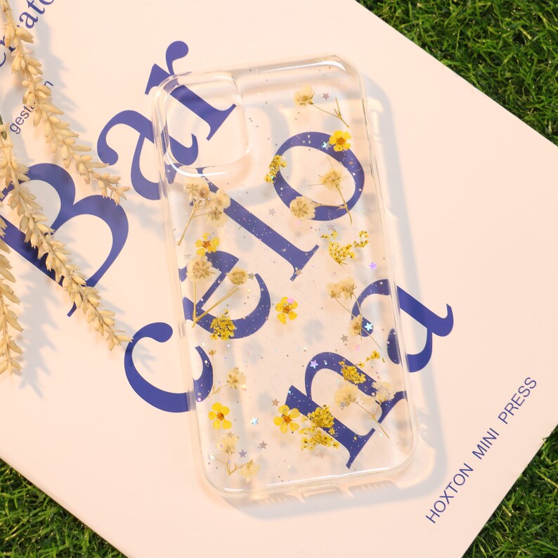 May include: A clear phone case with a blue "Hello" design and pressed yellow flowers and silver glitter.