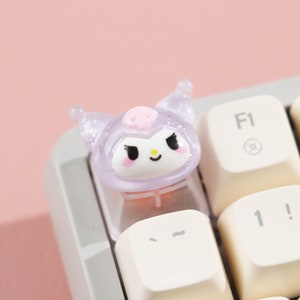 Puede incluir: Un tap&oacute;n de tecla de color morado y rosa con una cara blanca y ojos negros. El tap&oacute;n de tecla tiene forma de personaje de dibujos animados con orejas y capucha. El tap&oacute;n de tecla est&aacute; en un teclado blanco.