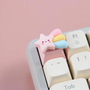 May include: A pink star-shaped keycap with a rainbow tail and a smiling face. The keycap is on a white keyboard with a pink key.