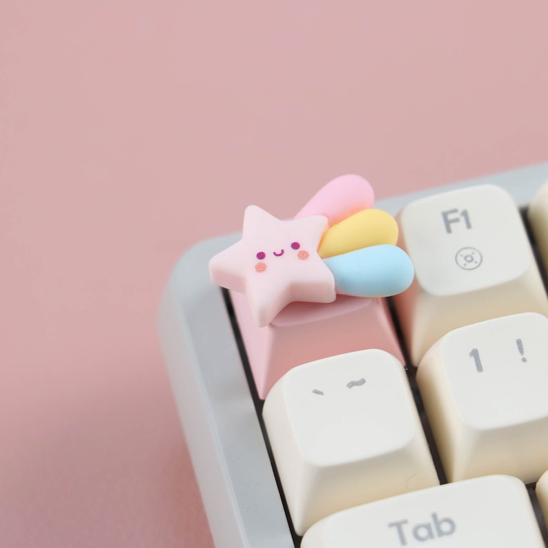 May include: A pink star-shaped keycap with a rainbow tail and a smiling face. The keycap is on a white keyboard with a pink key.