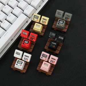 May include: Six pairs of miniature gaming consoles on small wooden stands. The consoles are in various colors, including red, white, black, pink, yellow, and gray.
