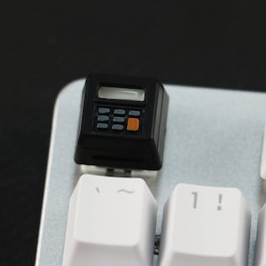 May include: A black calculator keycap with an orange button and a small display screen. The keycap is on a silver keyboard with white keys labeled with the symbols '~' and '1!'