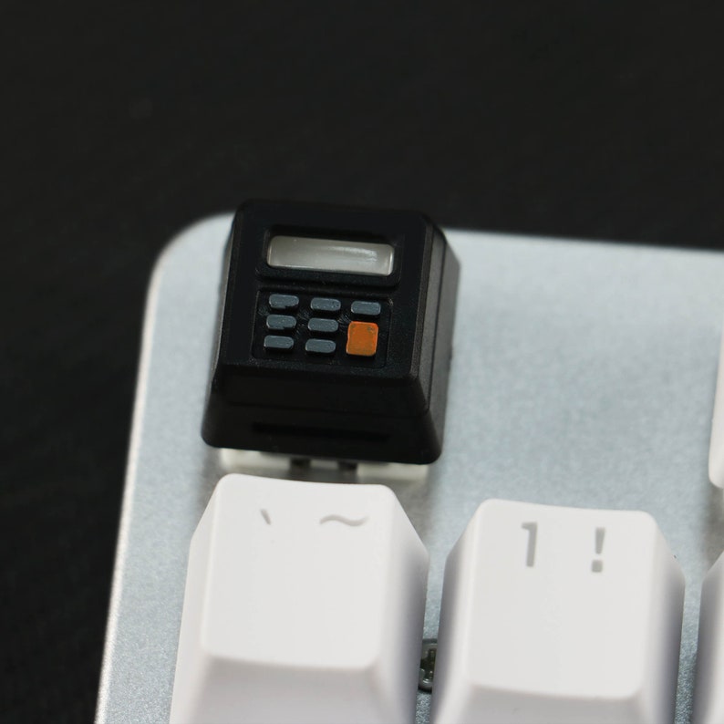 May include: A black calculator keycap with an orange button and a small display screen. The keycap is on a silver keyboard with white keys labeled with the symbols '~' and '1!'