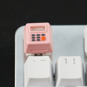 May include: A pink plastic keycap shaped like a calculator with a black display and gray buttons. The keycap is sitting on a white keyboard with the keys labeled "~" and "1!"