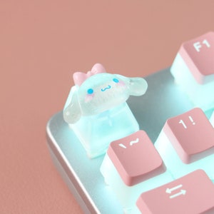 Puede incluir: Un tap&oacute;n de tecla de color azul claro y rosa con un lazo en la parte superior. El tap&oacute;n de tecla tiene forma de perro de dibujos animados con una cara blanca y ojos azules. El tap&oacute;n de tecla est&aacute; sentado en un teclado rosa y blanco.