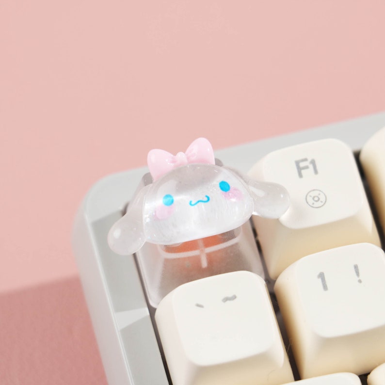 Puede incluir: Un tap&oacute;n de tecla de teclado transparente de pl&aacute;stico con un lazo rosa y una cara de personaje de dibujos animados. El personaje tiene ojos azules y una nariz rosa. El tap&oacute;n de tecla est&aacute; en un teclado blanco.