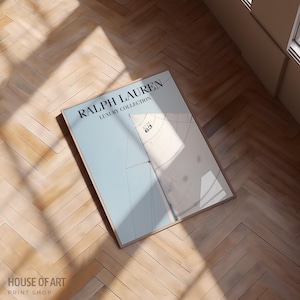 May include: A framed print featuring the text "RALPH LAUREN LUXURY COLLECTION" in black. The print has a light blue background with a white and beige design. The print is lying on a wooden floor.