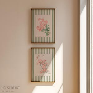 May include: Two framed floral prints. Each print features a pink floral arrangement on a cream background with a green striped border. The frames are a warm brown color. The prints are hanging on a cream-colored wall.