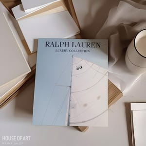May include: A light blue and white Ralph Lauren Luxury Collection book featuring a sailboat image. The book is open, revealing a partial view of a sailboat sail and mast. The image is on a white surface with other white and beige items.