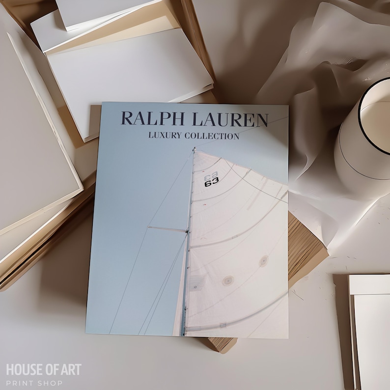 May include: A light blue and white Ralph Lauren Luxury Collection book featuring a sailboat image. The book is open, revealing a partial view of a sailboat sail and mast. The image is on a white surface with other white and beige items.