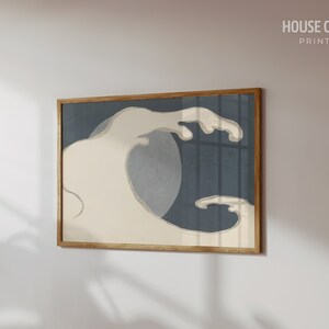 May include: A framed print of a stylized wave in shades of blue and white. The wave is depicted in a minimalist style, with a single, large crest and a smaller, trailing wave. The print is framed in a natural wood frame.