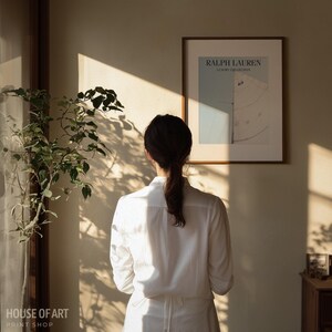 May include: A woman in a white shirt stands facing a framed poster with the text "RALPH LAUREN LUXURY COLLECTION". The poster has a light blue background with a white and gray design. A leafy plant is visible to the left.