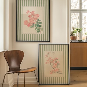 May include: Two framed floral prints with black frames. The prints feature pink flowers and green leaves on a cream background. The frames have a green and white striped border. A wooden chair sits in front of the prints.