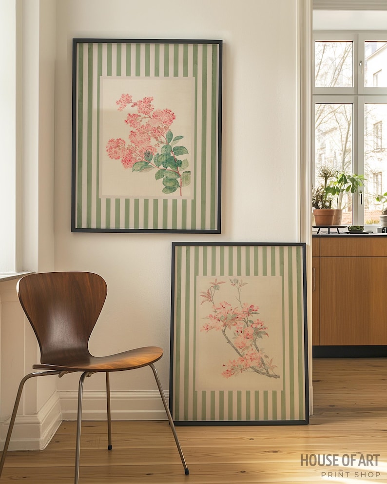 May include: Two framed floral prints with black frames. The prints feature pink flowers and green leaves on a cream background. The frames have a green and white striped border. A wooden chair sits in front of the prints.