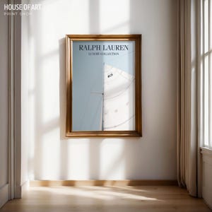 May include: A framed print featuring a sailboat with a white sail against a light blue sky. The print is in a gold-colored wooden frame and includes the text "RALPH LAUREN LUXURY COLLECTION". The print is hanging on a white wall.